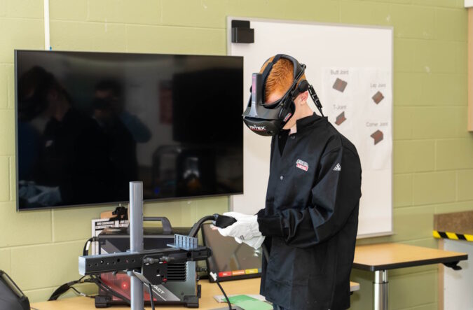 A man standing beside a large TV wearing a virtual reality headset and holding a controller.