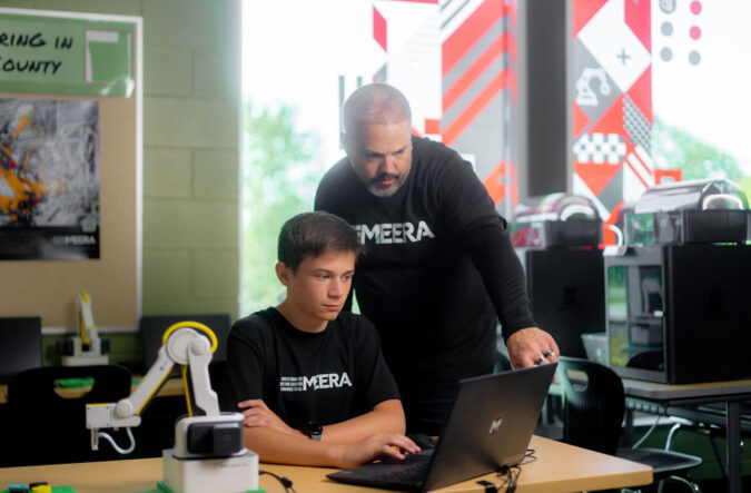 A student seated at a desk with an instructor standing behind him and pointing to the student's laptop screen.