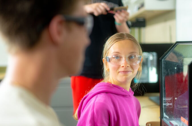 A student wearing safety glasses standing in front of a 3-D printer and looking at another student wearing safety glasses.