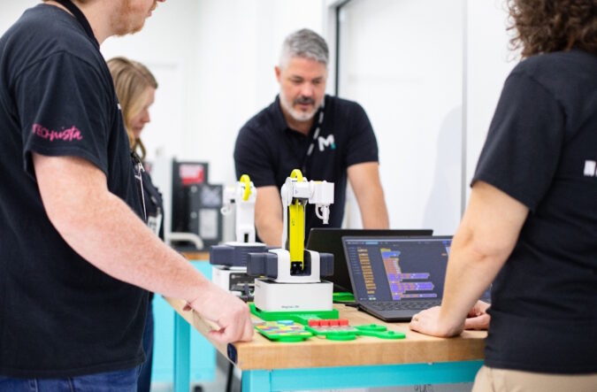 Four people gathered around a table with laptops and robotic arms on it.